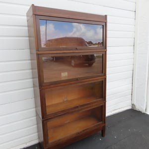 May include: A tall, four-tier wooden bookcase with a dark brown finish. Each of the four shelves features a clear glass front panel that lifts and slides upward. The unit stands on four short, straight legs and has a flat top surface.