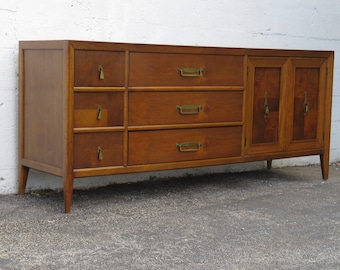 May include: A mid-century modern dresser with six drawers and two cabinet doors. The dresser is made of wood and has brass hardware. The dresser is in good condition and has a warm brown finish.