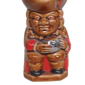 May include: A brown ceramic figurine of a man in a hat and red-trimmed coat, holding a silver object. The figurine has a stern expression and is standing upright. The base is rectangular.