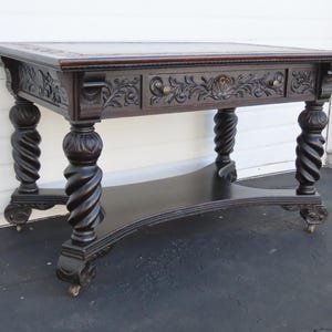 May include: A dark brown wooden desk featuring a rectangular top, a single drawer with intricate floral carvings, and two small round knobs. The desk is supported by four thick, spiraled legs that rest on small casters, with a curved lower shelf.