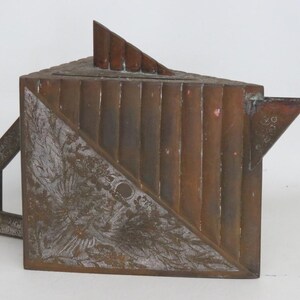 May include: A geometric copper teapot featuring a triangular body with vertical ribbed detailing on one side and an etched floral pattern on the other. The teapot includes a sharp, angular spout, a flat top lid with a fin, and a square handle.