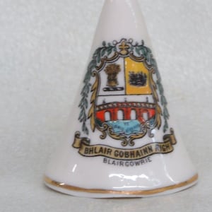 May include: White ceramic cone-shaped thimble with a gold trim. The thimble features a colorful coat of arms with a bridge, wheat, and a crown. The text "Blair Gobhainn Righ Blairgowrie" is printed on the thimble.