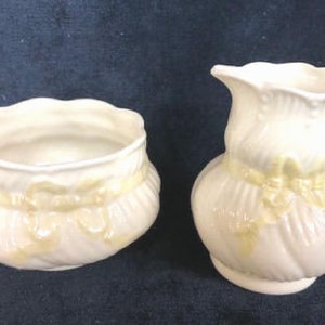 May include: A white porcelain creamer and sugar bowl set with yellow floral designs. The creamer has a handle and a spout. The sugar bowl is round and has a wide opening.