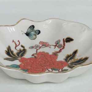 May include: A white ceramic bowl with a scalloped edge and a gold rim. The bowl features a floral design with a red flower, green leaves, and a blue butterfly.