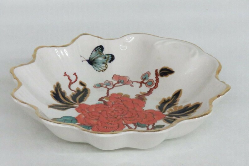May include: A white ceramic bowl with a scalloped edge and a gold rim. The bowl features a floral design with a red flower, green leaves, and a blue butterfly.