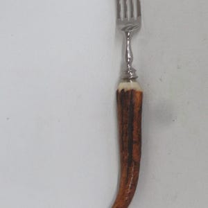 May include: A silver-toned metal fork featuring a four-tined head and a curved, textured handle made of brown and cream-colored antler material. The metal neck connects the tines to the organic, tapered handle, creating a distinct contrast in texture.
