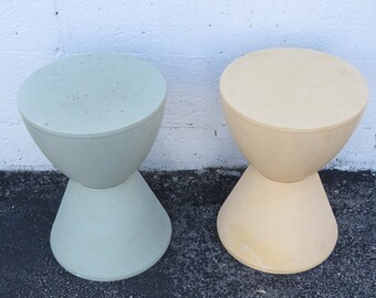 May include: Two hourglass-shaped stools, one in a light green color and the other in a light yellow color. The stools are made of plastic and have a smooth, rounded top.