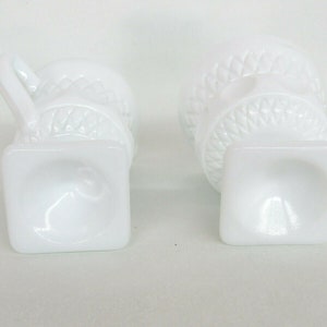 May include: Two white milk glass cups with a raised design. The cups have a square base and a round top. The cups are empty.