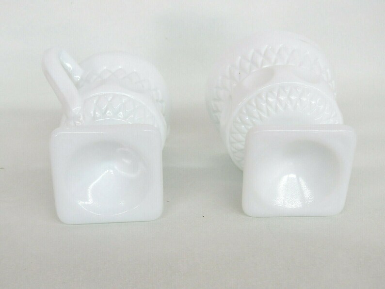May include: Two white milk glass cups with a raised design. The cups have a square base and a round top. The cups are empty.
