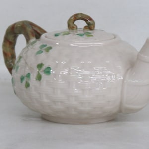 May include: A white ceramic teapot with a green shamrock design and a brown handle. The teapot has a woven pattern and is decorated with green shamrocks.