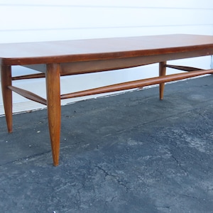 May include: A mid-century modern coffee table with a rectangular top and tapered legs. The table is made of wood with a warm brown finish. It features a lower shelf and a stretcher bar for added support. The table measures approximately 48 inches long.