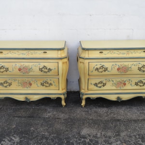 May include: Two matching antique yellow dressers with two drawers each. The dressers have a curved design with floral and scroll details. The trim is a dark teal color. The tops of the dressers are a light yellow color.