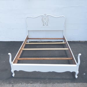 May include: White painted bed frame with a decorative headboard. The headboard features an ornate design with a central figure-eight shape. The frame has curved legs and a wooden slat support system.