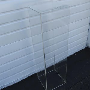 May include: A clear acrylic display stand with a rectangular shape. The stand is tall and slender, with a hollow interior and a solid base. The clear material allows for full visibility of items placed on the stand.