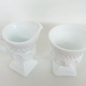 May include: A set of two white milk glass creamer and sugar bowl with a raised, embossed design. The creamer has a handle and a spout.