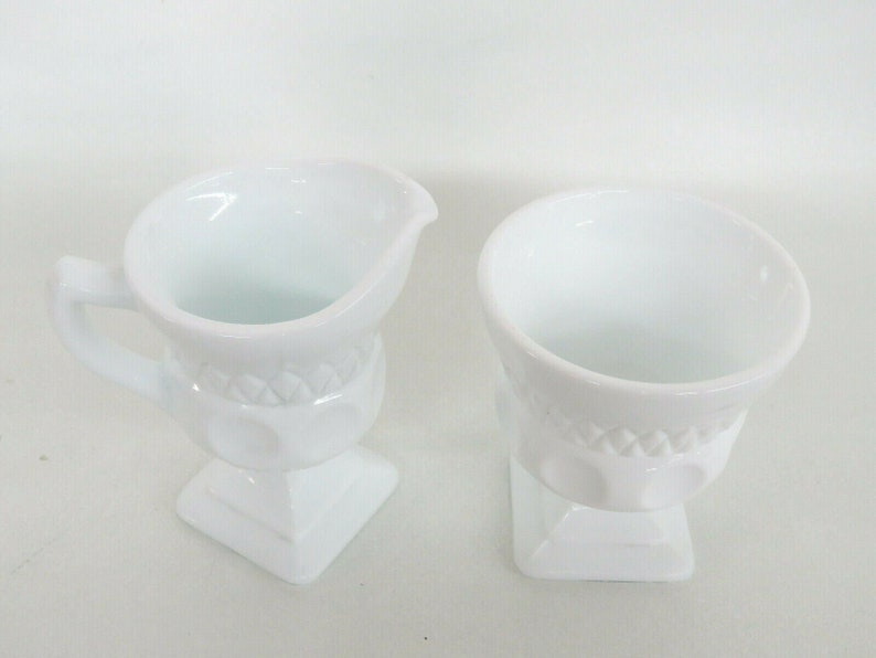 May include: A set of two white milk glass creamer and sugar bowl with a raised, embossed design. The creamer has a handle and a spout.