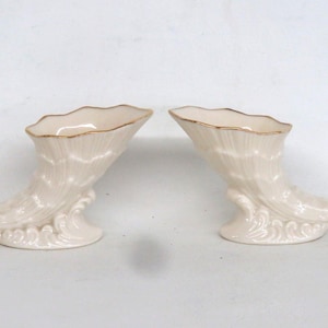 May include: A pair of cream-colored ceramic cornucopia vases featuring a ribbed shell-like texture and a gold-painted rim. Each vessel rests on a base sculpted with wave-like patterns, creating a symmetrical display of two identical horn-shaped containers.