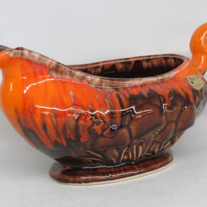 May include: A ceramic gravy boat with a vibrant orange and brown glaze. The gravy boat features a curved spout, a looped handle, and a textured exterior. The base is oval-shaped, and the overall design is reminiscent of a bird.