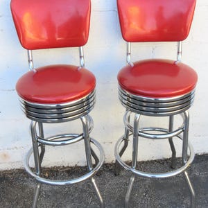 May include: Two retro-style bar stools with red seats and backs. The seats are round, and the backs are rectangular. The stools have chrome-colored metal frames with a footrest and four legs. The stools are set against a white wall.