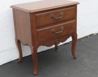 May include: A wooden nightstand with two drawers and brass handles. The nightstand has a curved apron and legs. The wood is a warm brown color. The nightstand is approximately 24 inches tall, 20 inches wide, and 16 inches deep.