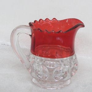 May include: A clear glass creamer with a red glass top. The creamer has a scalloped rim and a clear glass handle. The bottom of the creamer has a textured design.