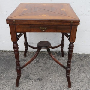 May include: A square, antique wooden table with a dark brown finish. The table features a decorative inlaid top, a drawer with a brass handle, and four ornate legs. The table's height is approximately 28 inches.