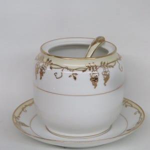 May include: A white porcelain sugar bowl with a matching saucer and spoon. The bowl and saucer have gold trim and a gold grape vine design. The spoon rests inside the bowl. The set is on a white background.