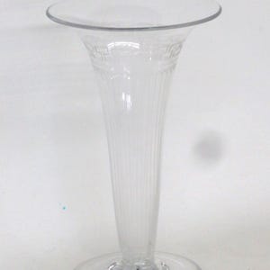 May include: A clear glass vase with a flared top and a round base. The vase has a long, slender body with vertical ridges and a decorative pattern near the top. The vase is designed to hold flowers.