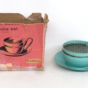 May include: A vintage turquoise cup and saucer smoke set with a metal grate inside the cup. The set includes a matching saucer and a copper-colored handle. The original box is also included, with the words "cup & saucer smoke set" printed on it.