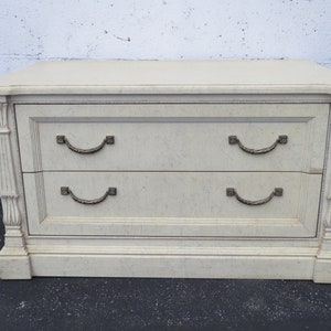 May include: A white two-drawer dresser with ornate details and brass handles. The dresser has a textured surface and a slightly distressed look.
