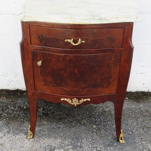 May include: A small, antique wooden nightstand with a marble top. The nightstand has a single drawer and a small door, both with decorative gold hardware. The wood has a rich, dark brown finish with a patterned grain, and the legs have gold accents.