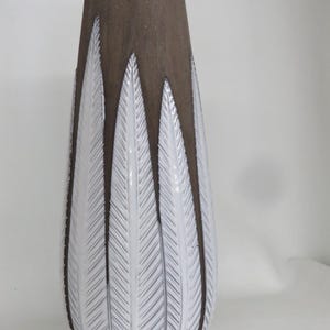 May include: A tall, decorative vase with a brown upper section and a white lower section. The white section features a repeating feather-like pattern. The vase has a smooth, glossy finish and a tapered shape, suitable for home decor.