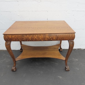 May include: A vintage wooden coffee table with a square top and a lower shelf. The table has ornate, curved legs and a warm, natural wood finish. The table measures approximately 30 inches long, 30 inches wide, and 20 inches high.