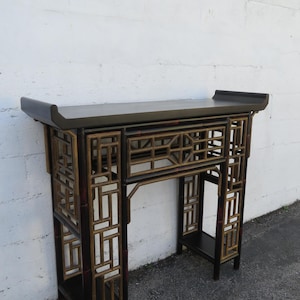 May include: A dark brown and gold bamboo console table with a rectangular top and open geometric fretwork design. The table has a shelf and is against a white wall.