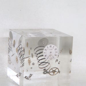 May include: A clear resin cube encases various watch parts, including gears, springs, and a watch face. The cube is transparent, allowing a full view of the intricate details of the watch components suspended within. The watch parts are in shades of silver and gold.