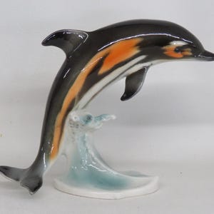 May include: A ceramic dolphin figurine in a leaping pose, with a black and orange body, white accents, and a light blue wave base. The dolphin is approximately 6 inches tall, a decorative item.