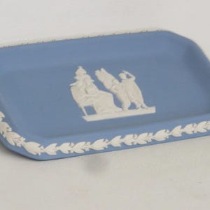 May include: A rectangular, light blue ceramic tray with a white relief design. The tray features a white border with a leaf pattern and a central relief of classical figures. A decorative piece.