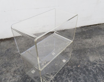 May include: A clear acrylic two-tiered shelf unit. The unit has a rectangular shape with rounded corners and a smooth, transparent surface. The shelf is designed for display or storage.