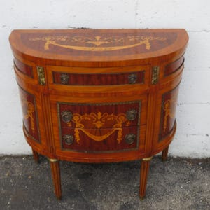 May include: A half-moon shaped wooden cabinet with two drawers and a curved top. The cabinet is made of a warm-toned wood with decorative inlaid patterns and brass hardware. It stands on three slender legs.