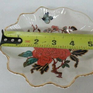May include: A white porcelain trinket dish with a scalloped edge and a gold trim. The dish features a floral design with red, blue, and green flowers and leaves. A butterfly is painted on the dish.