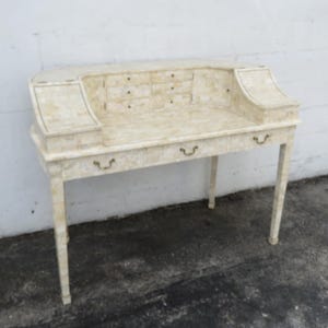 May include: A vintage-style desk with a cream-colored finish. It features a curved top with small drawers and three pull-out drawers below. The desk has slender legs and brass-toned hardware. The desk is approximately 40 inches wide.