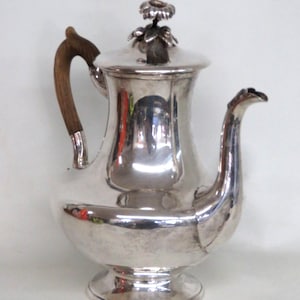 May include: A polished silver coffee pot featuring a curved wooden handle and a matching silver lid topped with a decorative floral finial. The vessel has a wide, rounded base that tapers toward the neck, a long curved spout, and a circular pedestal foot.