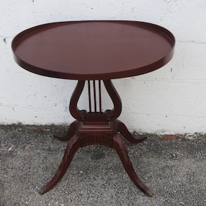 May include: A dark brown, antique side table with a round top and a unique lyre-shaped support. The table has three curved legs and stands approximately 28 inches tall. The table is made of wood and has a glossy finish.