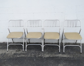May include: Four vintage folding chairs with silver metal frames and tan padded seats. The chairs are arranged in a row against a white textured wall. The chairs are designed with a vertical slat back.
