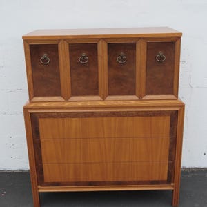 May include: A wooden chest of drawers featuring a top section with four cabinet doors, each equipped with a circular brass pull, and a bottom section with three horizontal drawers. The piece displays a mix of light and dark wood grain finishes.