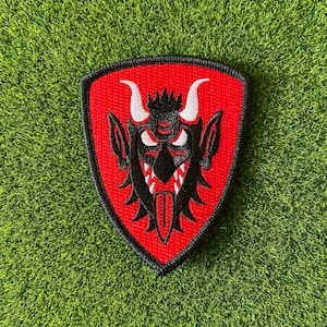 May include: A red, shield-shaped patch featuring a black devil face with white horns and teeth. The patch has a black border and is set against a green, textured background.