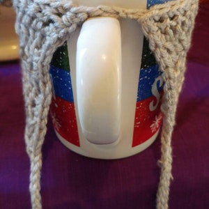 May include: A white ceramic mug with a colorful design and a gray crocheted cozy. The cozy has long ties that hang down on either side of the mug.