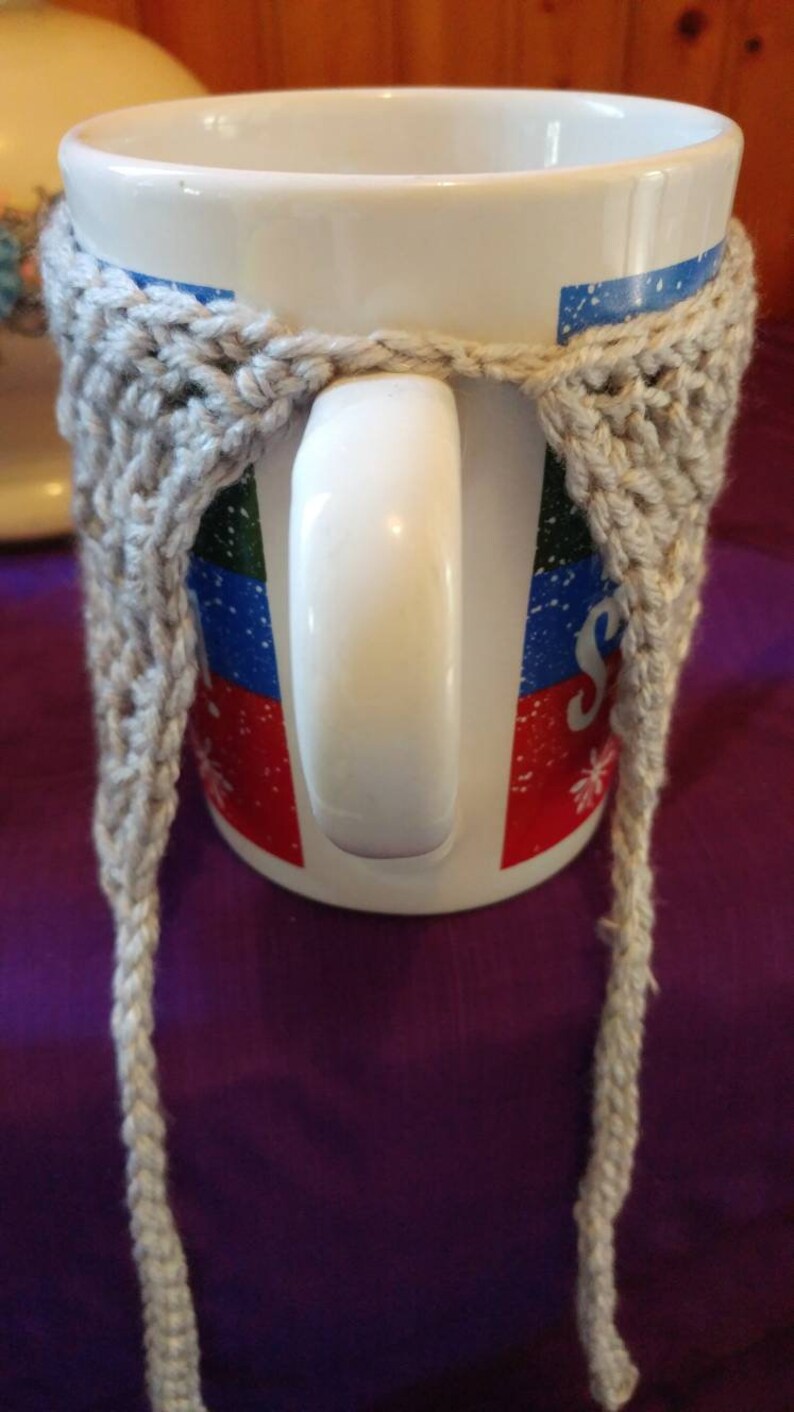May include: A white ceramic mug with a colorful design and a gray crocheted cozy. The cozy has long ties that hang down on either side of the mug.
