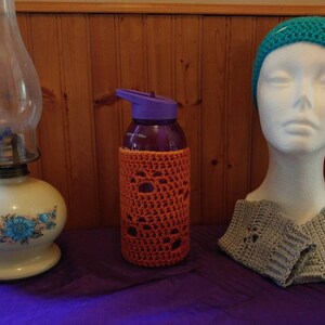 May include: A teal blue crocheted hat, a gray crocheted scarf, and a purple water bottle with an orange crocheted cover. The items are on a purple table.
