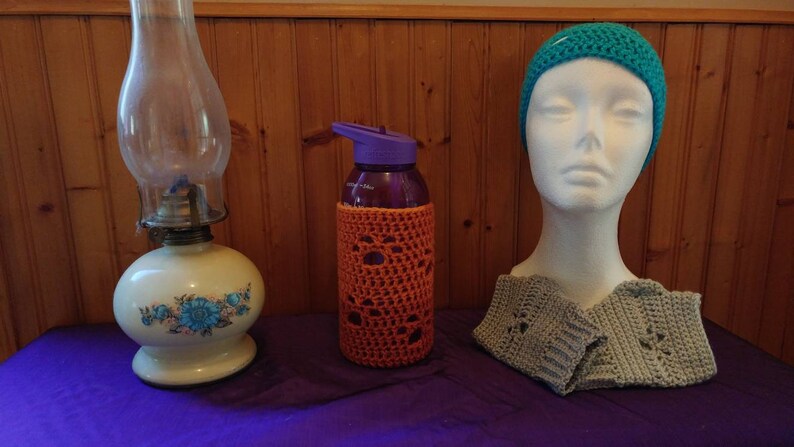 May include: A teal blue crocheted hat, a gray crocheted scarf, and a purple water bottle with an orange crocheted cover. The items are on a purple table.